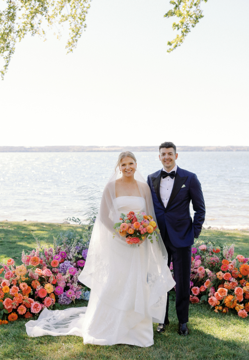 Bride and groom stand next to each other at ceremony altar, surrounded by color florals and with the lake behind them.
