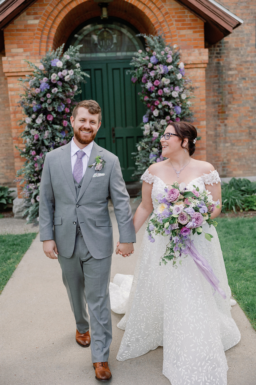Bride and groom holding hands and smiling at each other outside a stone church, walking past a floral archway of purple and lavender roses on the church's green doors.