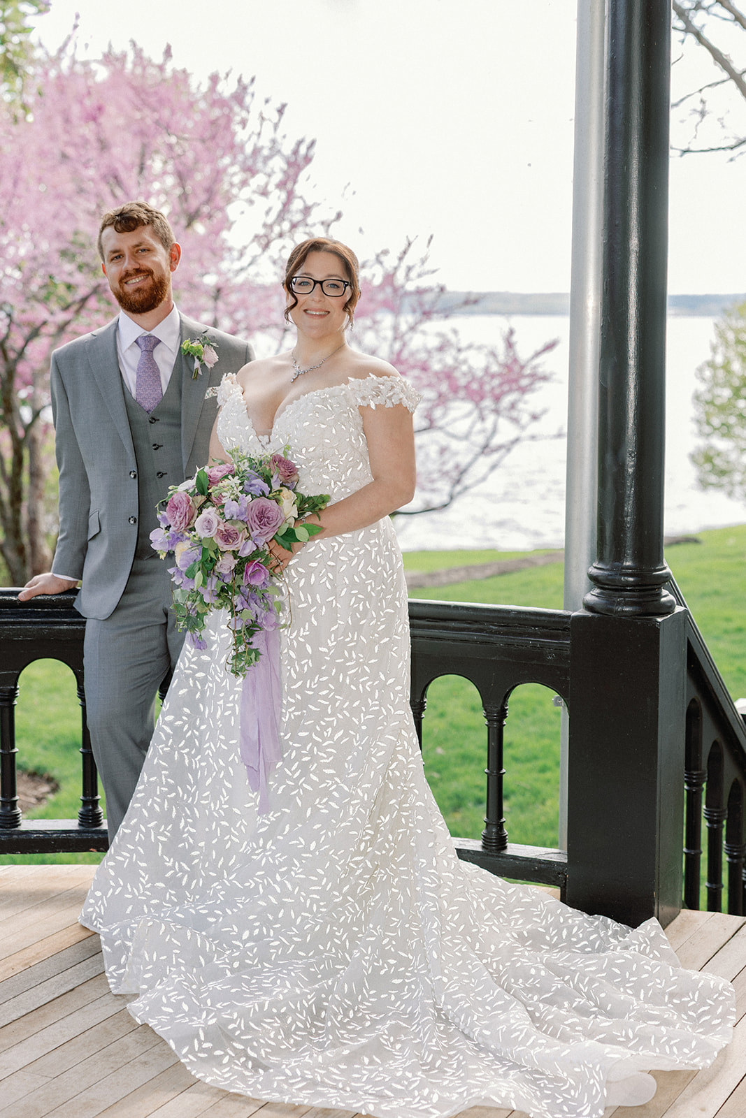 Bride and groom standing together on a black wrought-iron balcony overlooking the lake, the bride holding a cascading bouquet of lavender and blush roses, with blooming pink redbud trees behind them.