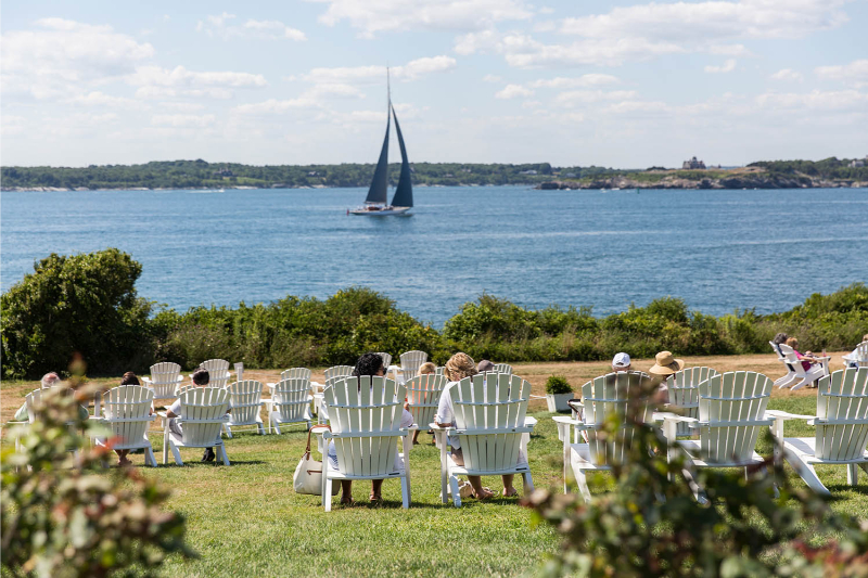 People sitting in lawn chairs at Castle Hill Inn overlooking the coast with a sailboat in the distant. 