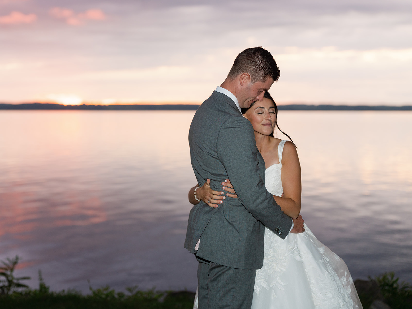 Bride and groom embrace with a lake behind them.