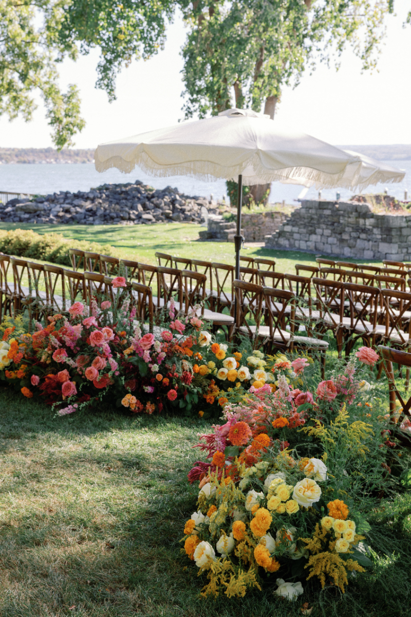 Ceremony setup at Inns of Aurora with wooden chairs, colorful aisle florals, and cream scalloped umbrellas.