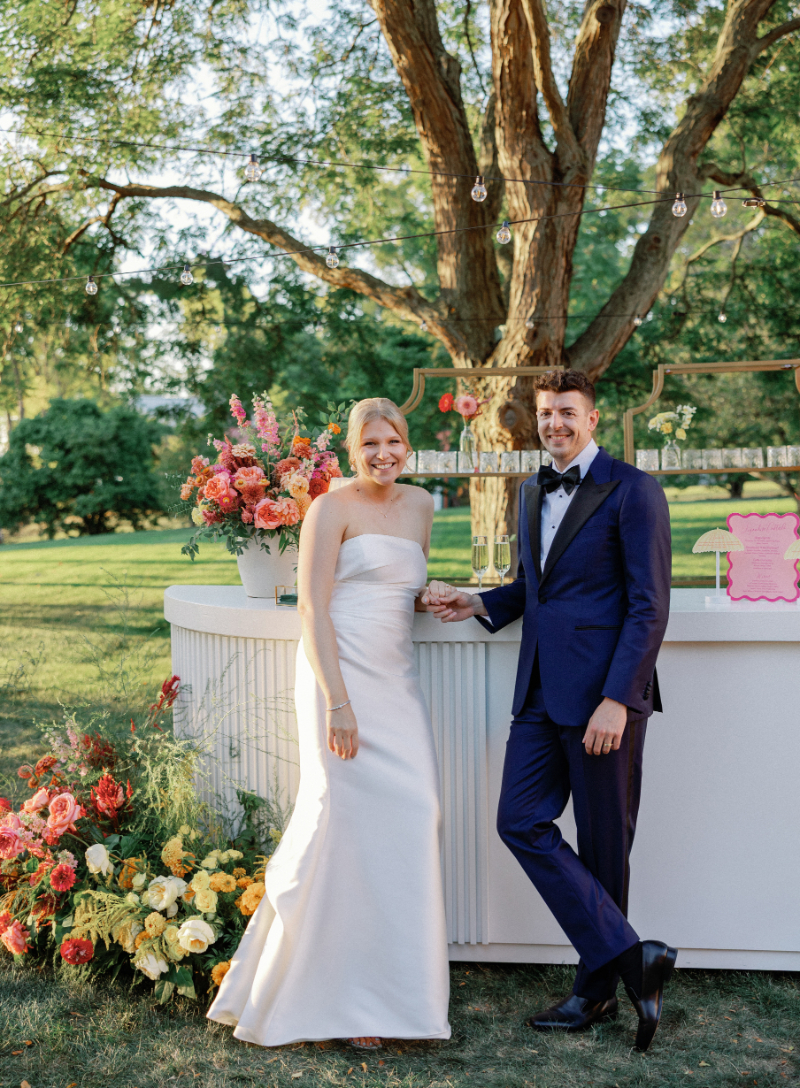 Bride and groom stand next to each other in front of cocktail hour bar, surrounded by Slim Aarons inspired decor.