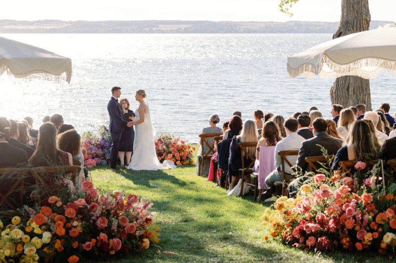 Bride and groom exchange vows with a lake view behind them, guests watch as they sit under umbrellas.