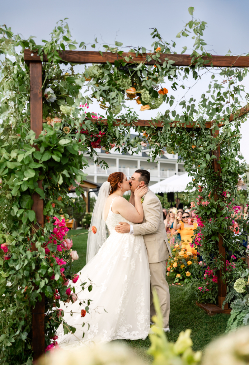 Bride and groom share first kiss under floral-filled chuppah.