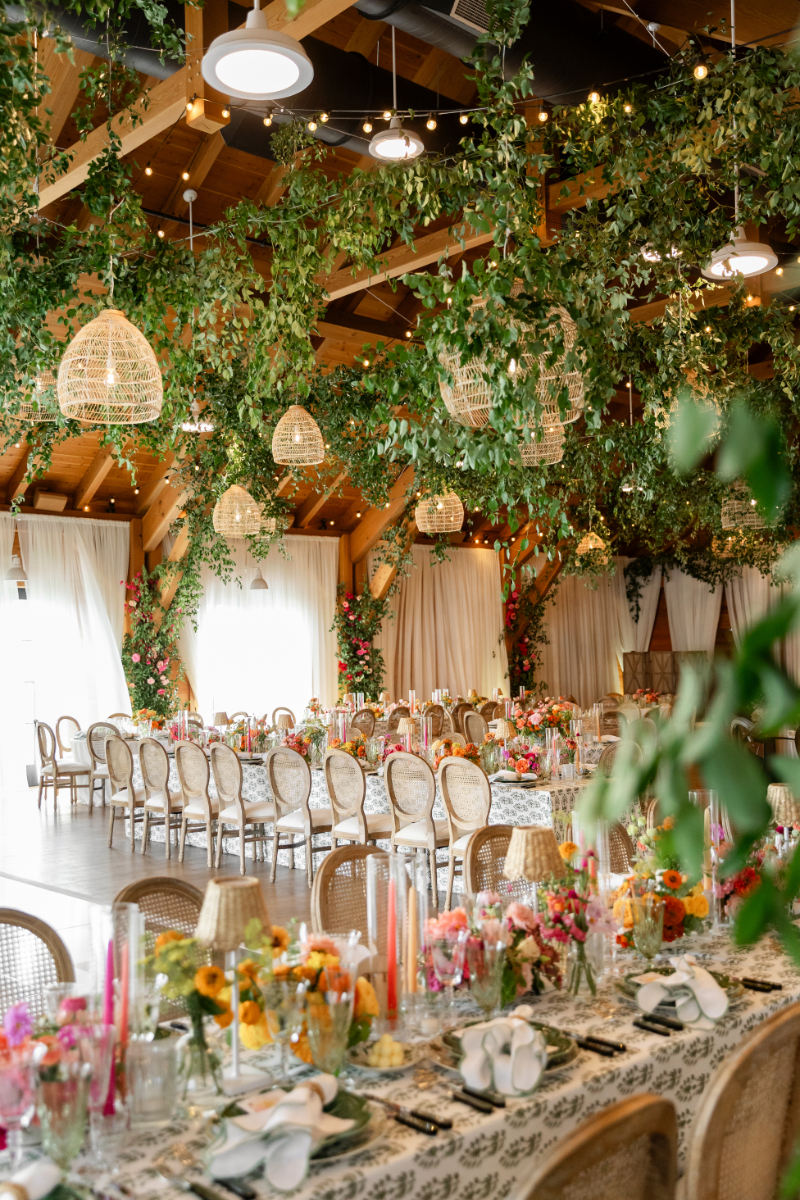 Colorful summer camp wedding reception at The Lake House on Canandaigua with rattan lanterns and chairs, greenery hanging from the ceiling and printed linen tablecloths.