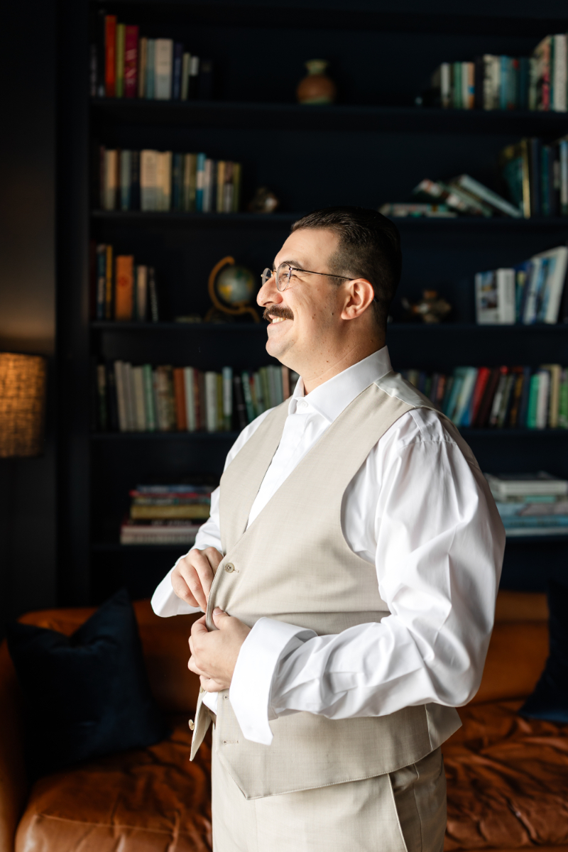 Groom buttons his vest as he looks out the window smiling.