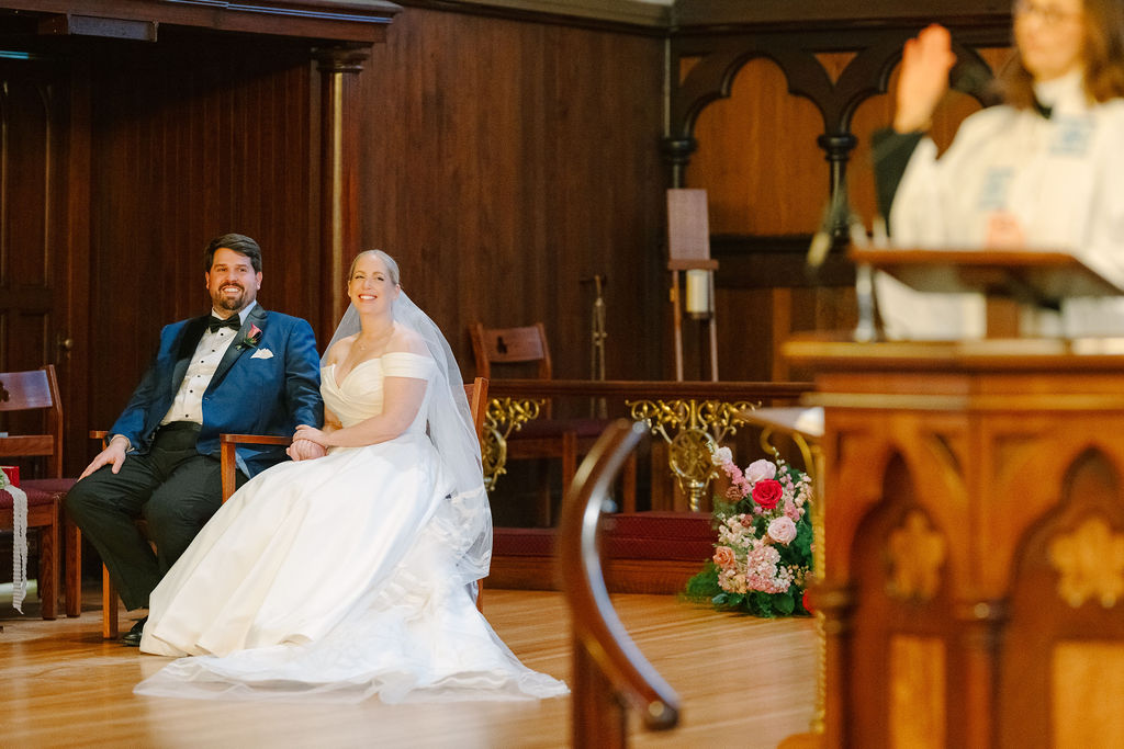 Bride and groom sit next to each other as pastor speaks.