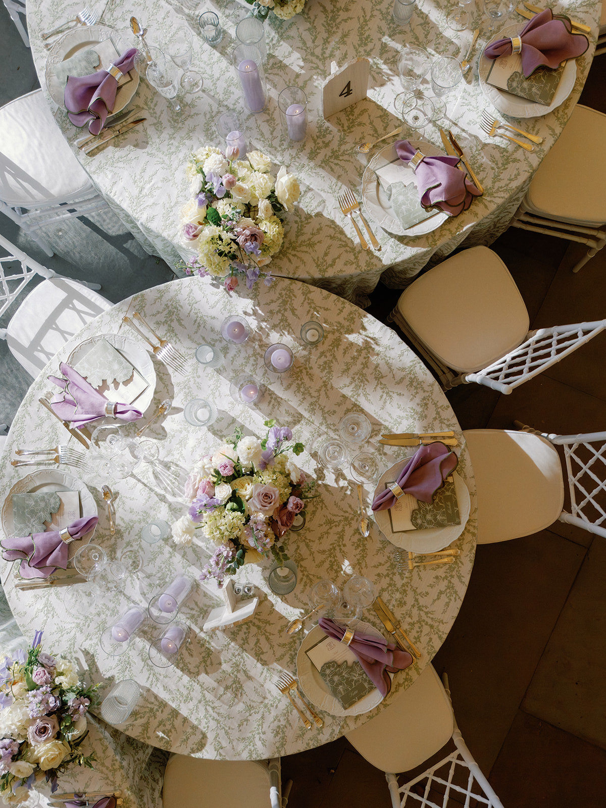 Overhead view of two round reception tables set with sage fern-print linens, lavender napkins in gold rings, gold flatware, and low ivory and lavender floral centerpieces, flanked by white chairs.