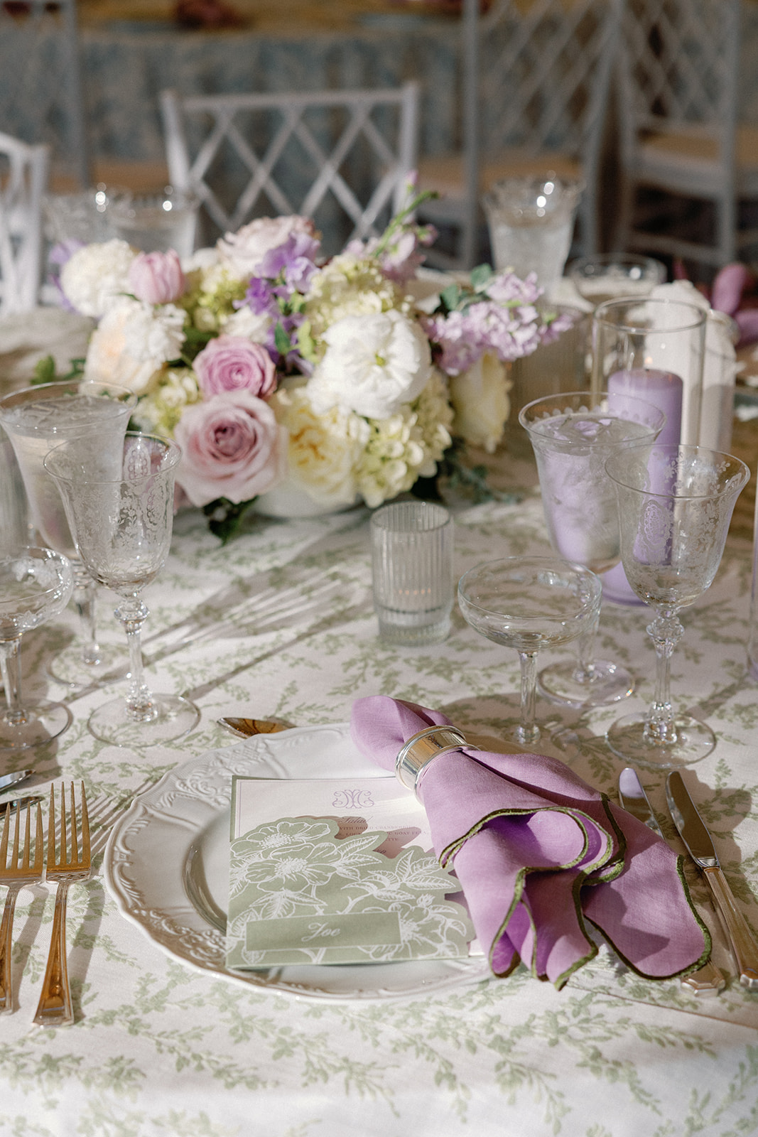 Close-up place setting with a lavender napkin, a green floral place card reading 'Joe,' gold flatware, and crystal glassware, with a floral centerpiece blurred in the background.
