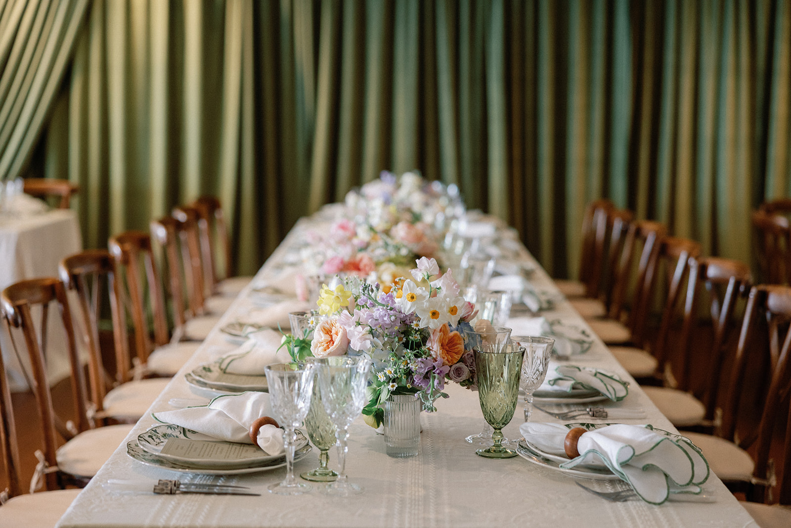 Long table set with a garland of pastel flowers running down the center, flanked by crystal and green glassware and green-patterned china, against a sage curtain backdrop.