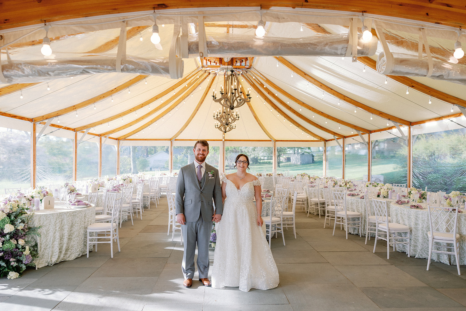 Bride and groom standing together holding hands inside the empty tented reception space before guests arrive, wooden chandeliers and string lights overhead.