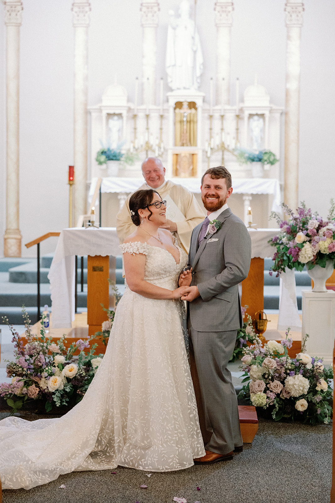 Bride and groom smiling and holding hands at the altar, with a priest in white vestments standing behind them beside pastel floral arrangements.
