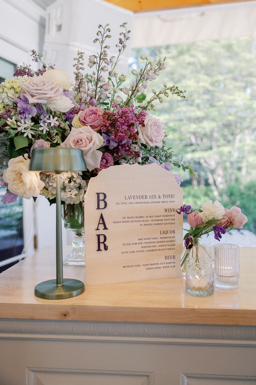 Close-up of a wooden bar sign reading 'Bar — Lavender Gin & Tonic' with a wine, liquor, and beer menu, beside a lush pink and lavender floral arrangement.