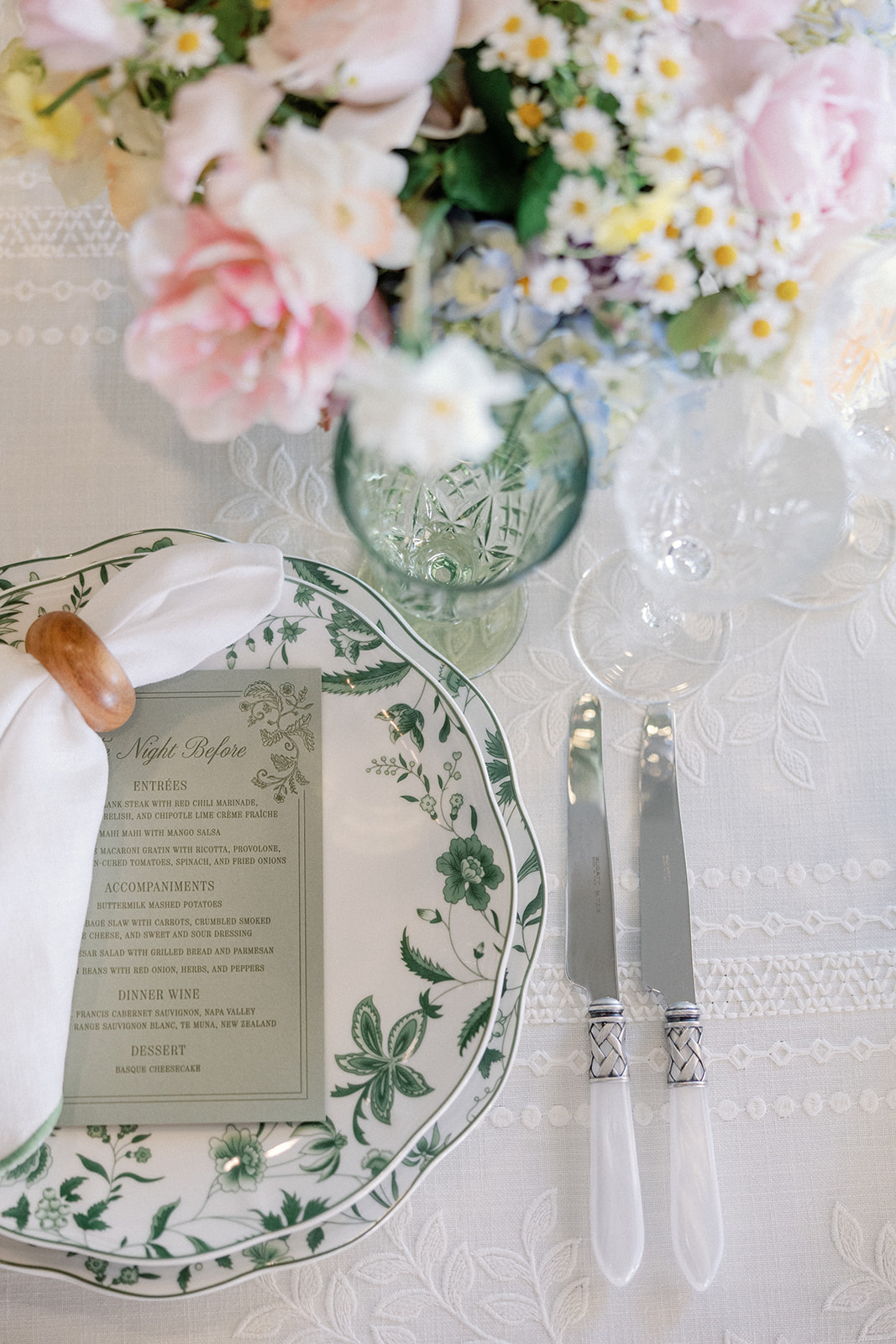 Overhead view of a place setting with a green floral menu card reading 'The Night Before' on patterned green china, a napkin in a wooden ring, and two pearl-handled knives, with blush and white flowers blurred above.