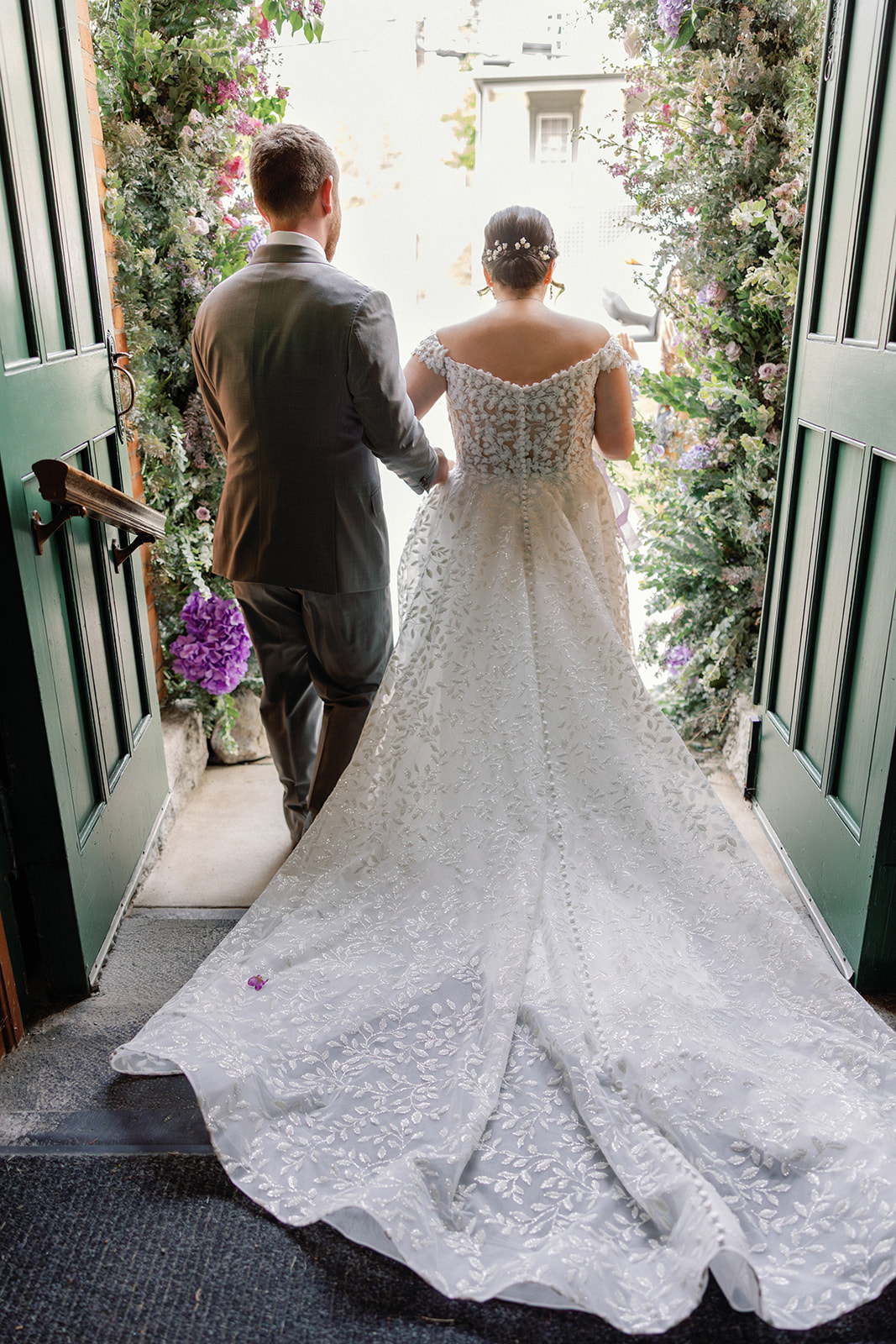 Back view of bride and groom walking out through open green doors framed by cascading floral garlands, her beaded gown trailing behind her.