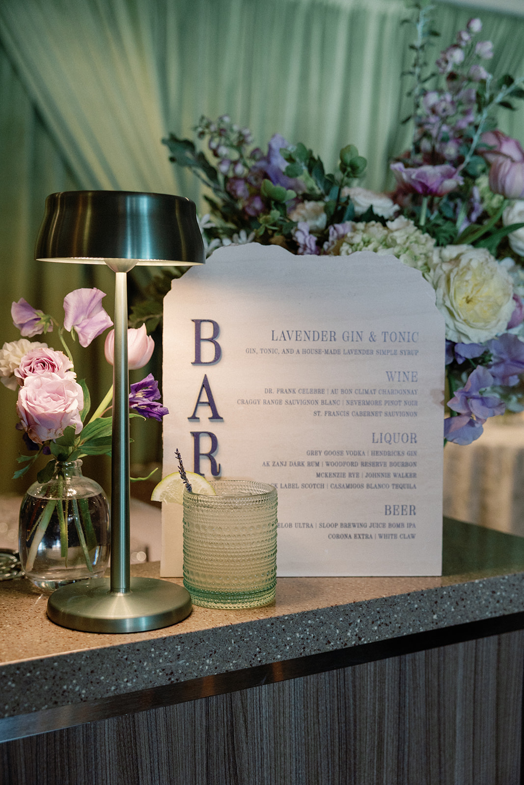 Close-up of a wooden bar sign reading 'Bar — Lavender Gin & Tonic' with a cocktail menu, beside a small vase of pink and lavender flowers and a garnished drink on a green draped curtain backdrop.