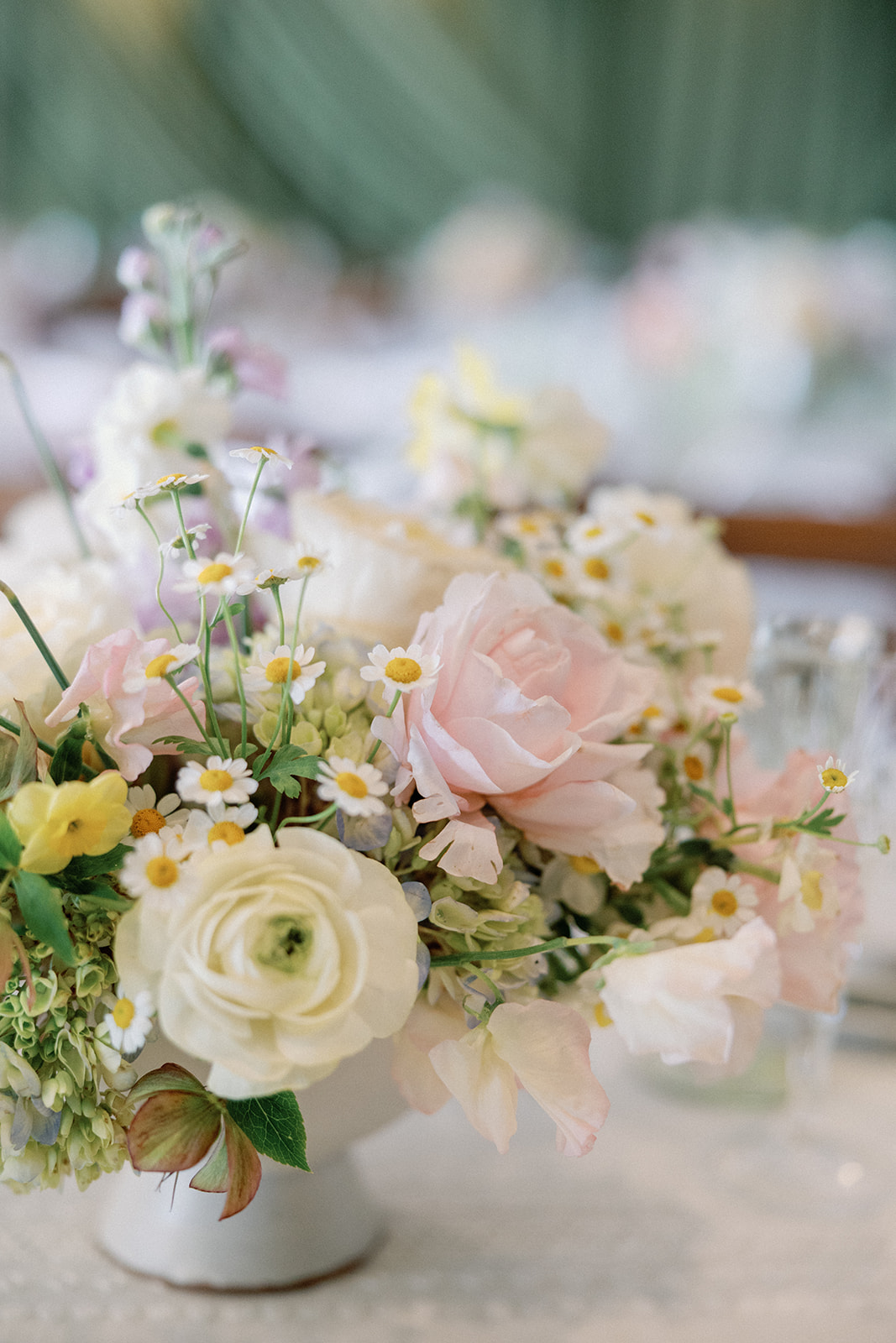 Close-up of a floral arrangement with blush garden roses, white ranunculus, chamomile daisies, and soft yellow blooms in a white vase.
