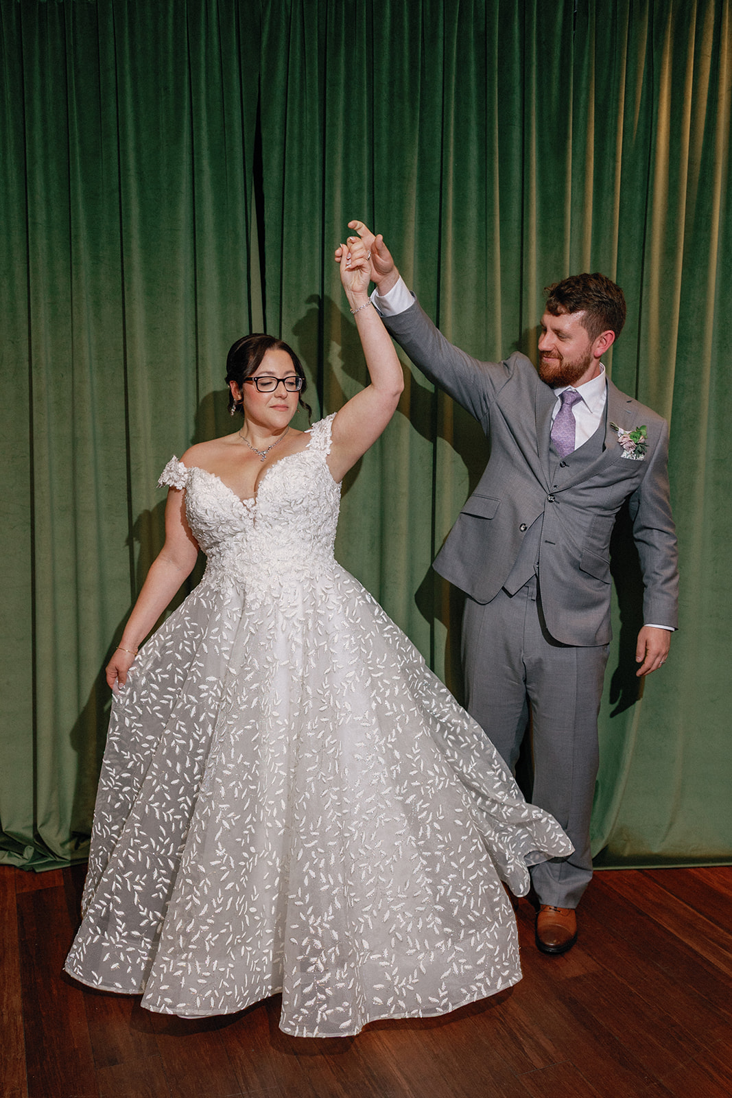 Bride and groom dancing together in front of a green velvet curtain, the groom twirling the bride as her beaded ball gown swirls around her.