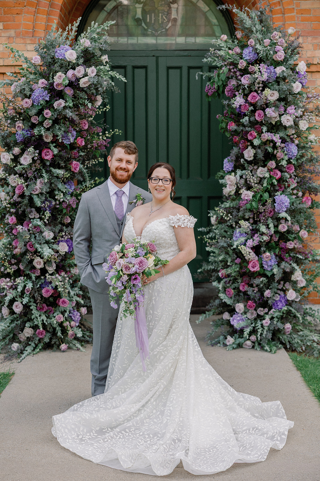 Bride and groom portrait standing together in front of green church doors framed by a lush arch of pink, purple, and lavender flowers.