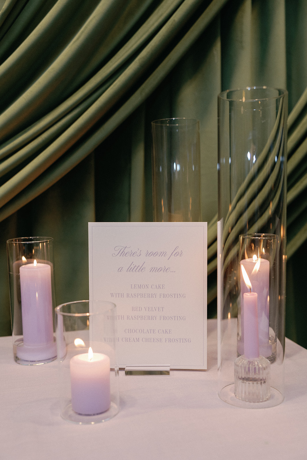 Dessert table detail with lavender pillar candles in glass cylinders and a sign reading 'There's room for a little more... Lemon cake with raspberry frosting, red velvet with raspberry frosting, chocolate cake with cream cheese frosting,' against a green velvet curtain.