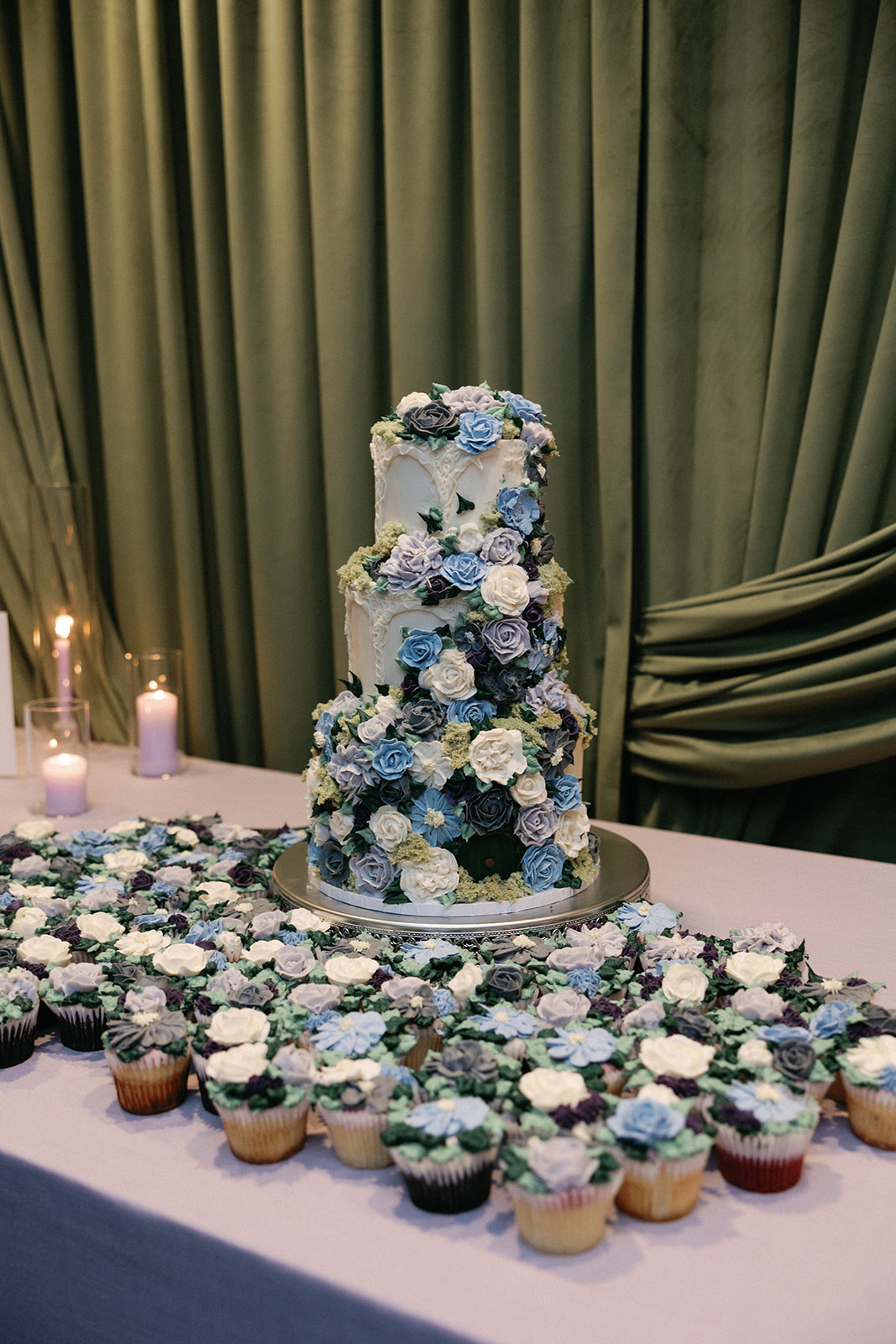 Three-tier wedding cake covered in blue, gray, and white buttercream flowers and greenery, surrounded by a spread of matching floral-topped cupcakes on a table draped in blue.