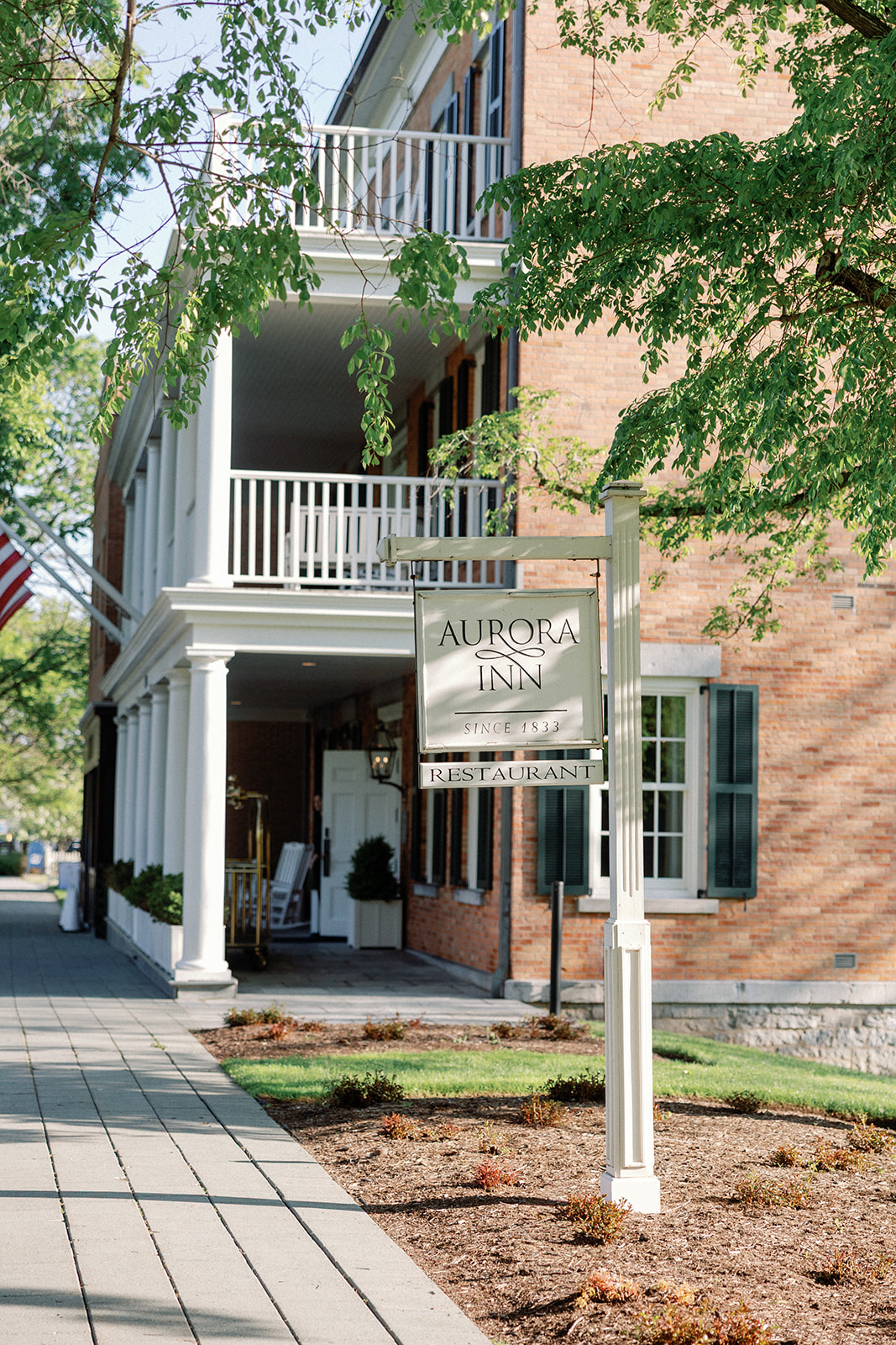 Exterior of the brick Aurora Inn building with white columns and balconies, framed by tree branches, with a hanging sign reading 'Aurora Inn Since 1833 Restaurant.'
