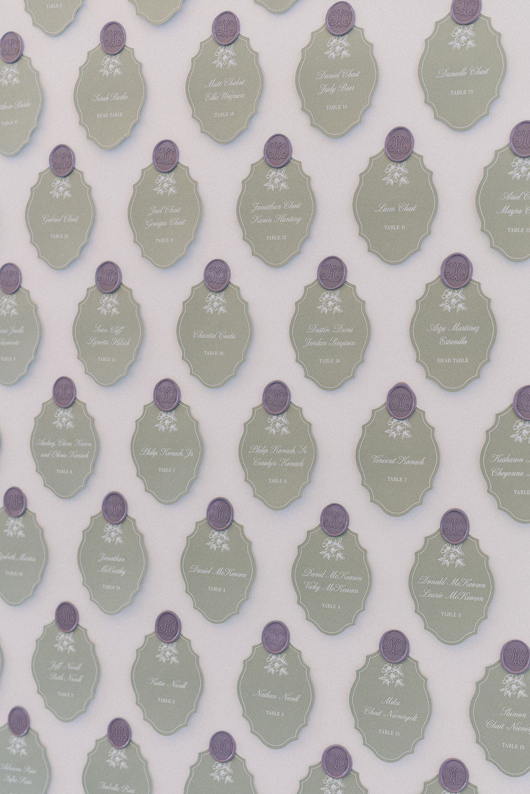 Close-up of an escort card display with rows of sage green oval cards topped with lavender wax seals, guest names and table numbers written in white script.