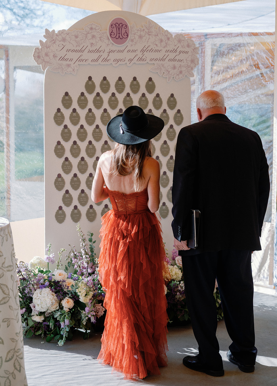 Back view of a wedding guest in a wide-brimmed black hat and rust ruffled gown standing beside an older man in a black suit, both looking at an escort card display topped with the quote 'I would rather spend one lifetime with you than face all the ages of this world alone.