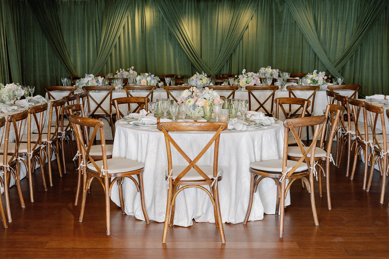 Round reception table set with crystal glassware and a low floral centerpiece in front of a draped sage green curtain, with cross-back wooden chairs and long feasting tables in the background.