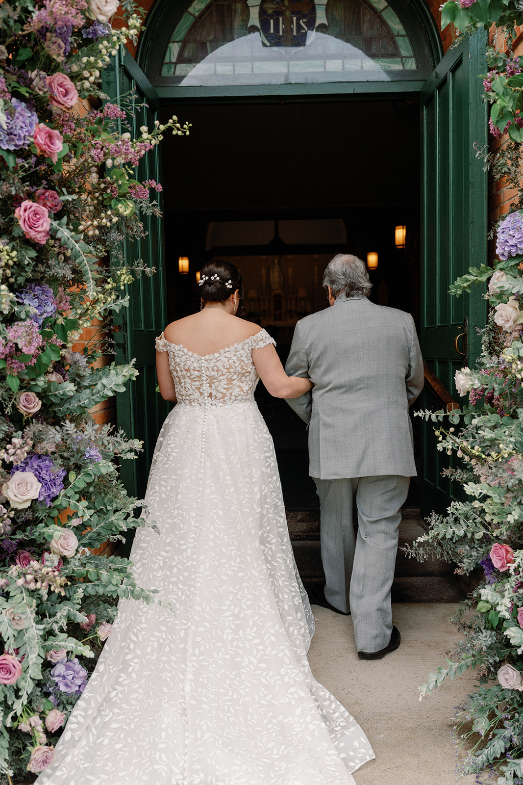 Back view of a bride and her father walking arm in arm into the church, framed by floral arrangements on either side of the open doorway.