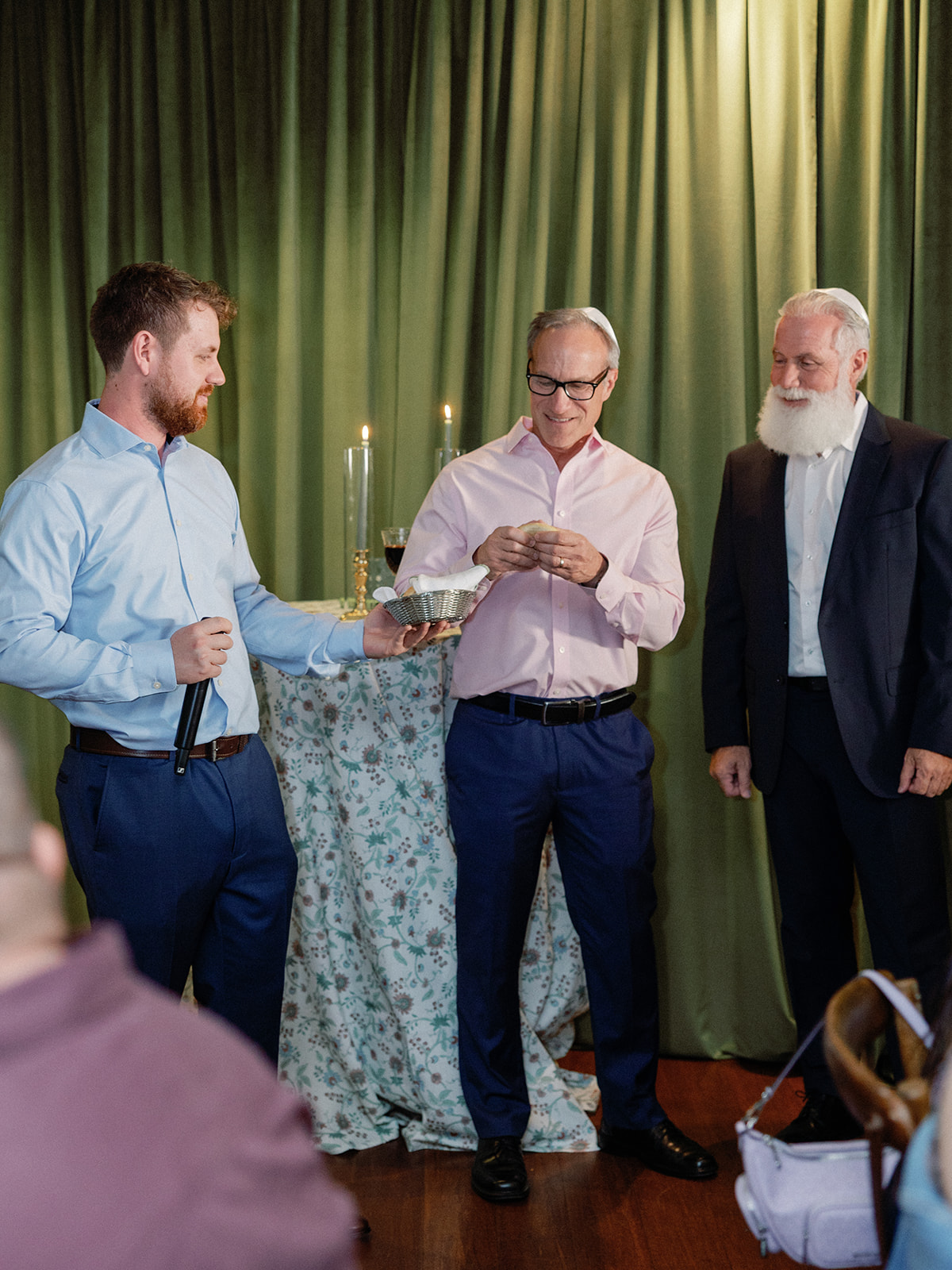 Three men standing before a green draped curtain and a table set with lit taper candles, one holding a microphone, another holding a small basket of bread during a blessing.