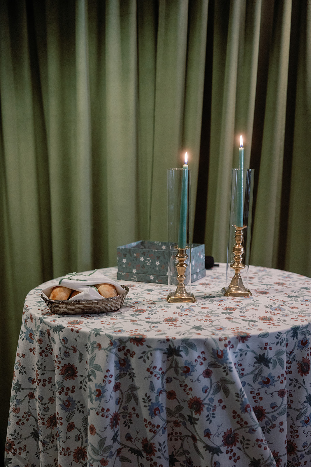 Round Shabbat table with a floral-print tablecloth, a basket of challah rolls wrapped in a napkin, a patterned box, and two tall teal taper candles burning in brass candlesticks, set against a draped sage green curtain.