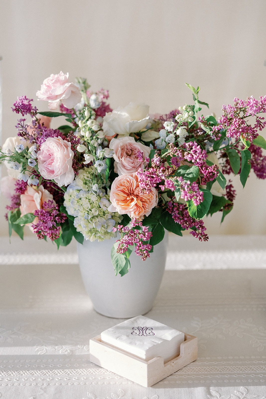 Close-up of a lush floral arrangement with blush and peach garden roses, purple lilac, white hydrangea, and delicate blue and white blooms in a pale gray vase, with a monogrammed napkin set in a wooden holder in front.