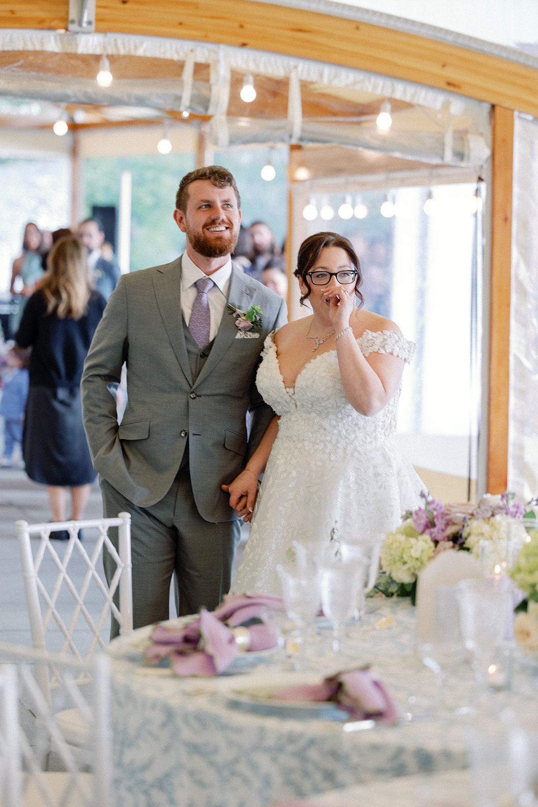Bride and groom standing together under the tent, the groom smiling and the bride with her hand raised to her mouth, visibly emotional, guests and a lavender-set table in the background.
