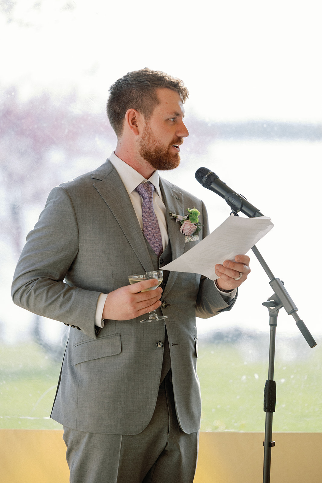 Groom giving a toast into a microphone, holding a glass of champagne and reading from a sheet of paper, with a blurred pink blossoming tree visible through the window behind him.