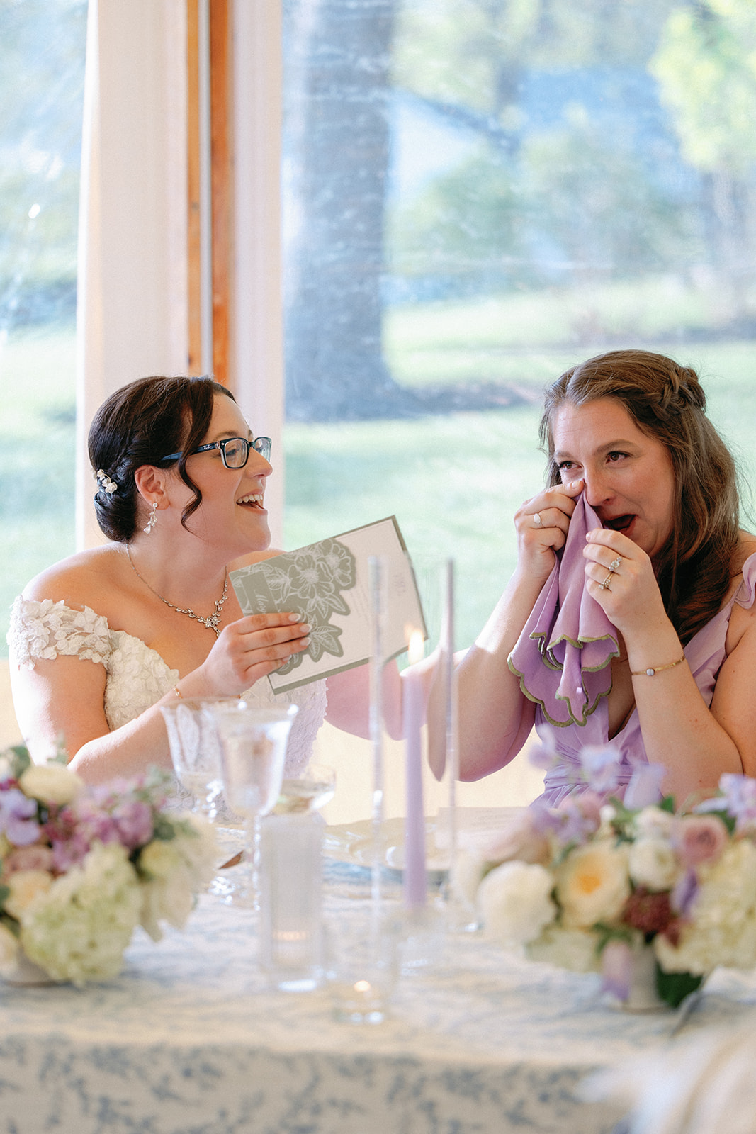 Bride laughing while showing a menu card to a bridesmaid in a lavender dress, who wipes away a tear with her napkin, both seated at a table with floral centerpieces and lit taper candles.