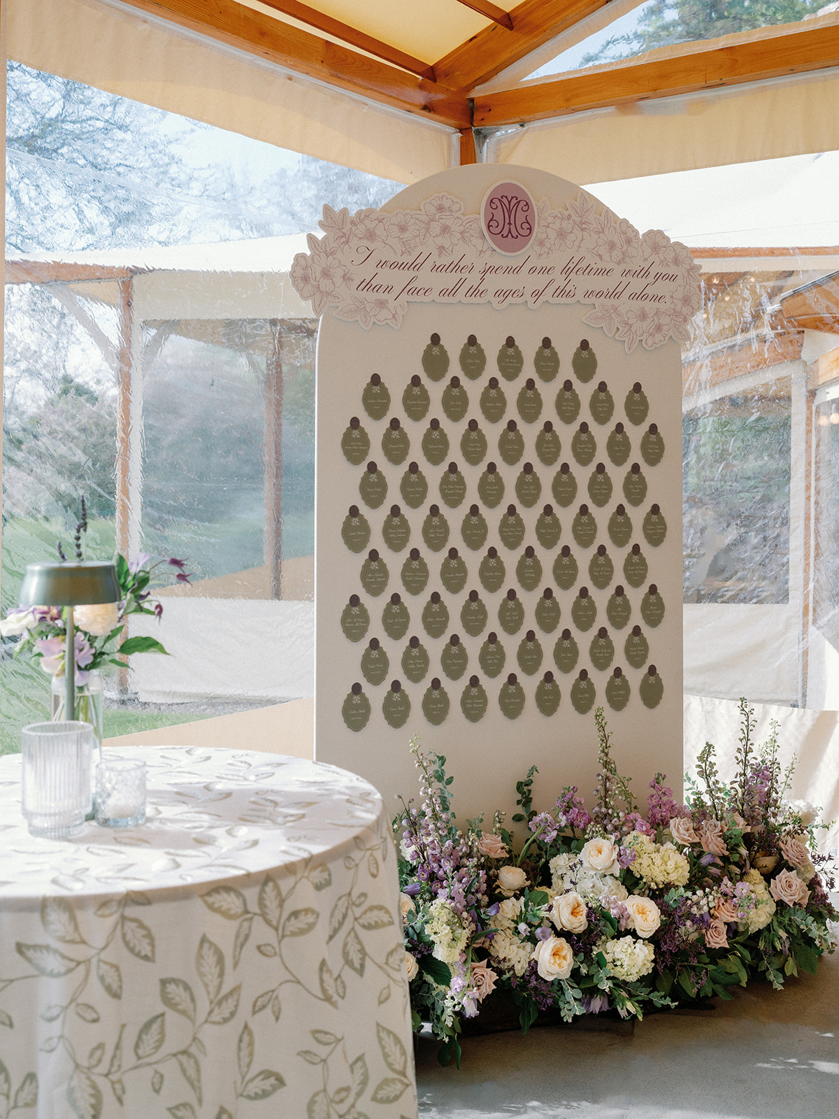 Wide view of the escort card display board under the tent, rows of sage cards topped with wax seals beneath the handwritten quote, with a floral arrangement at its base and a leaf-print table nearby.