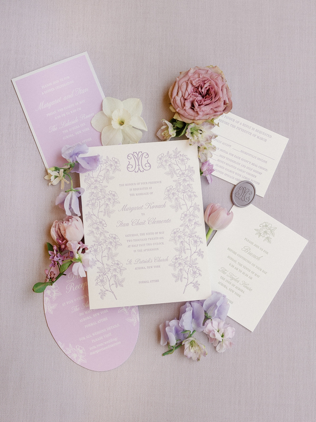 Wedding invitation suite flat lay with a floral-bordered invitation for Margaret Kovach and Itan Chait Clemente, a lavender reception card, a brunch card, a wax seal, and dried rose, tulip, and sweet pea blooms.