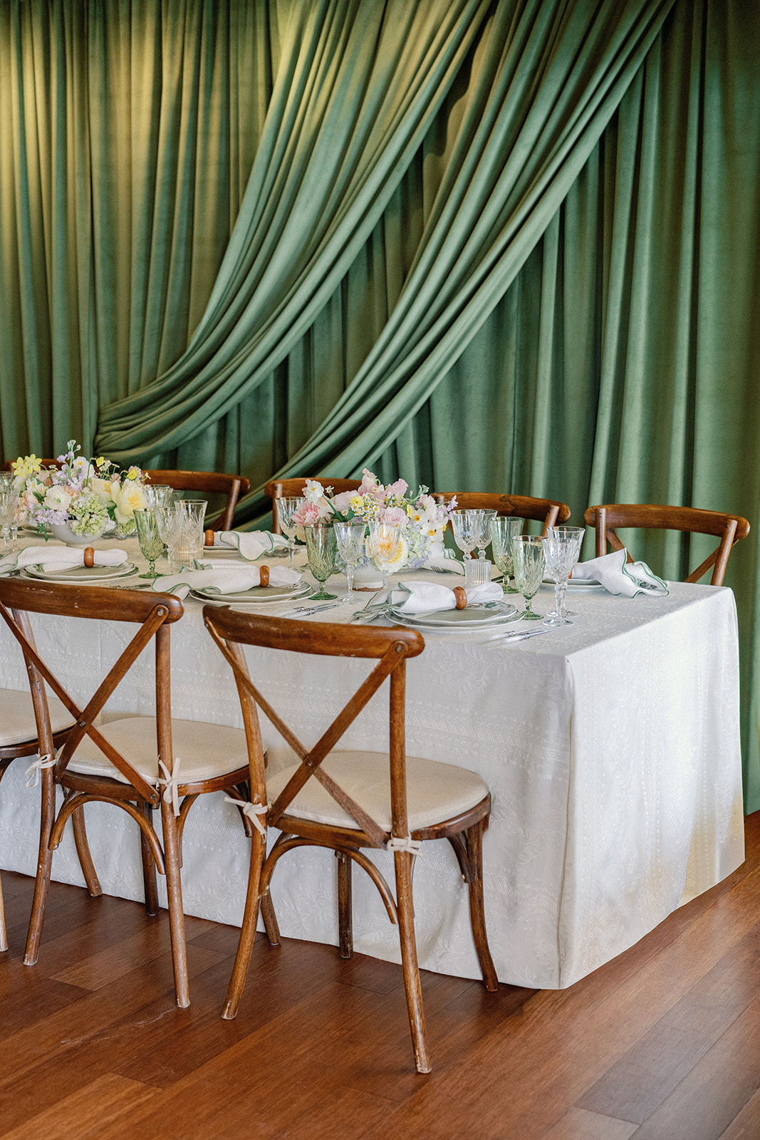 Long feasting table set with green and white china, green-rimmed glassware, and floral centerpieces against a pleated sage green curtain backdrop, wooden cross-back chairs in the foreground.
