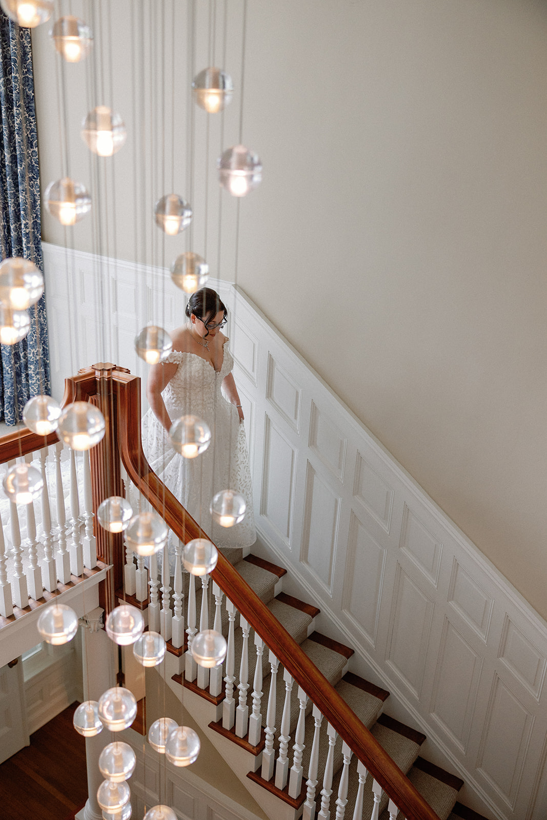 Overhead view of a bride in a lace off-the-shoulder gown descending a wood-paneled staircase, seen through a curtain of hanging glass bubble lights.