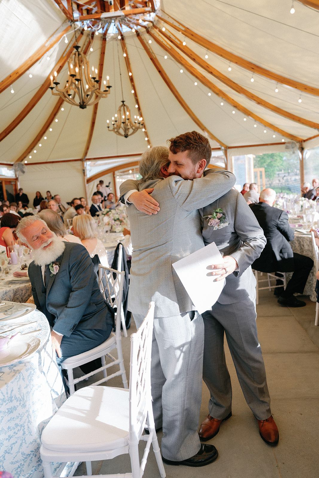 Groom embracing an older guest in a warm hug inside the tented reception, wooden chandeliers and string lights overhead and guests seated at floral-set tables in the background.