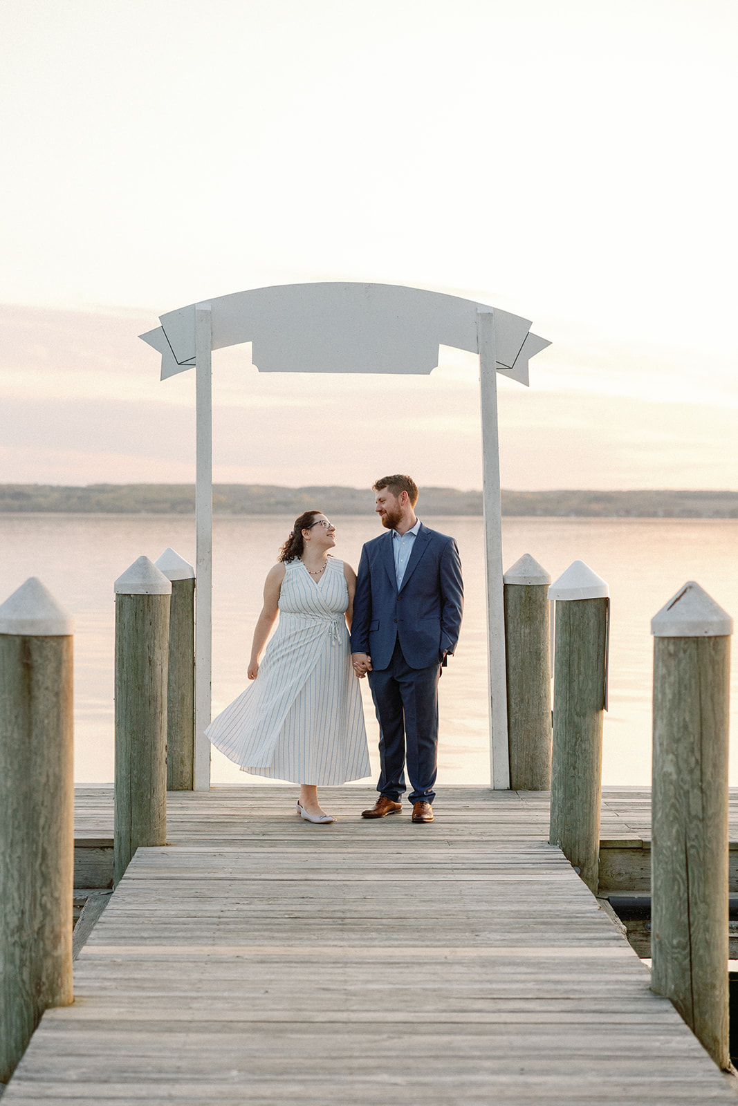 Bride and groom holding hands and smiling at each other under a white wooden arch at the end of a dock, with a lake and rolling hills glowing pink and orange at sunset behind them.