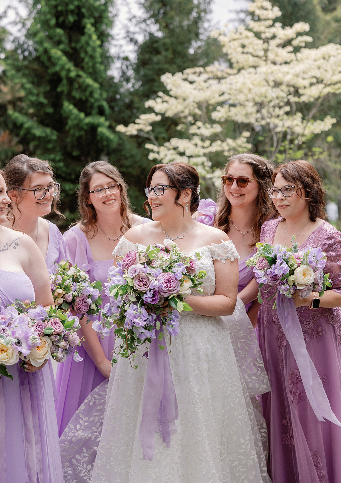 Bride surrounded by bridesmaids in lavender and dusty purple dresses, all smiling and holding bouquets, with a blooming white dogwood tree and evergreens behind them.