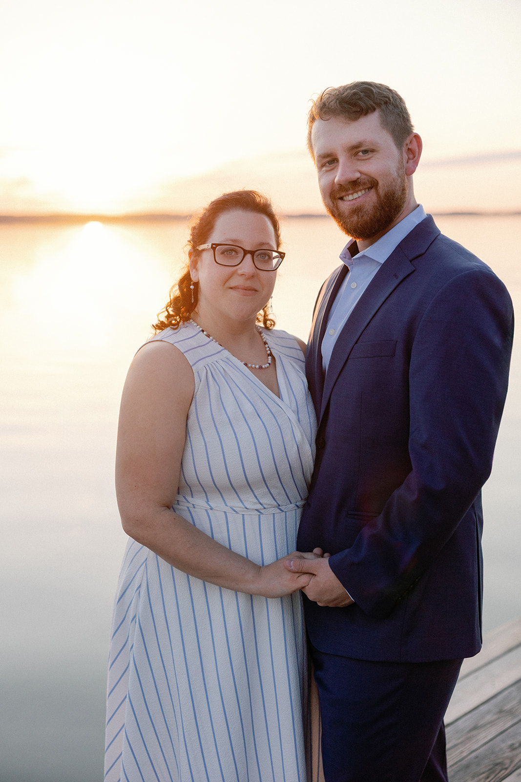 Close-up portrait of a bride and groom smiling at the camera with a pink and gold sunset over the water behind them.