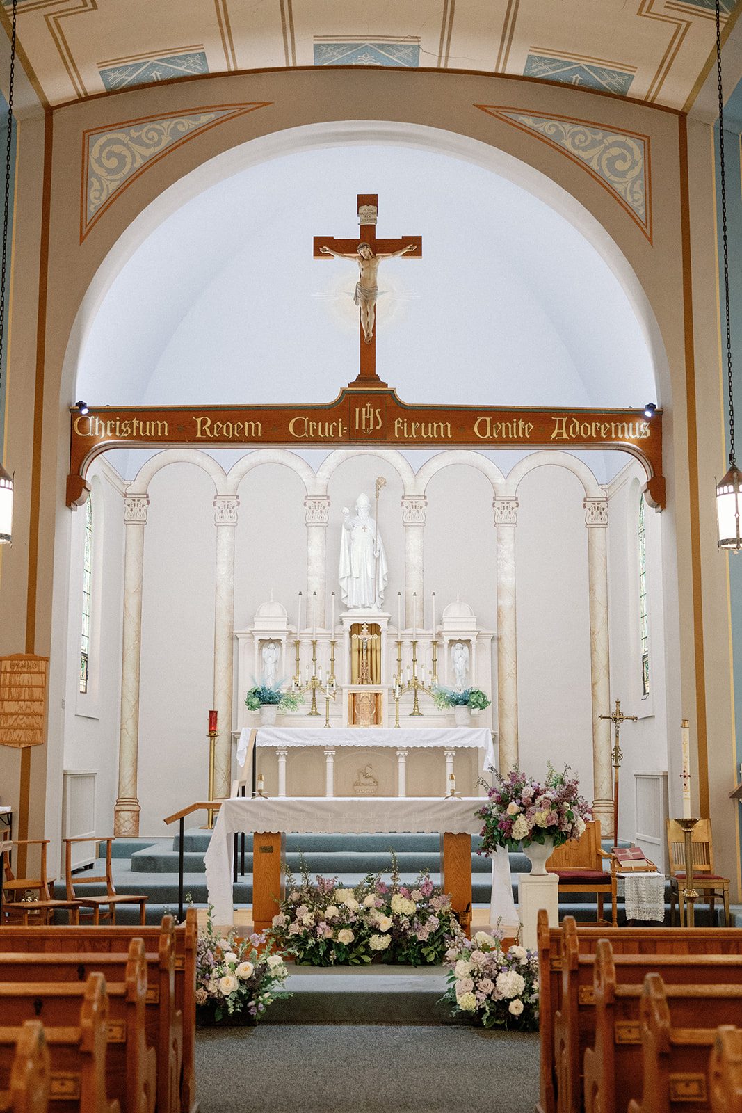 Ornate white church altar with columns, a statue, and a suspended crucifix above a Latin inscription, framed by lush pastel floral arrangements at its base.
