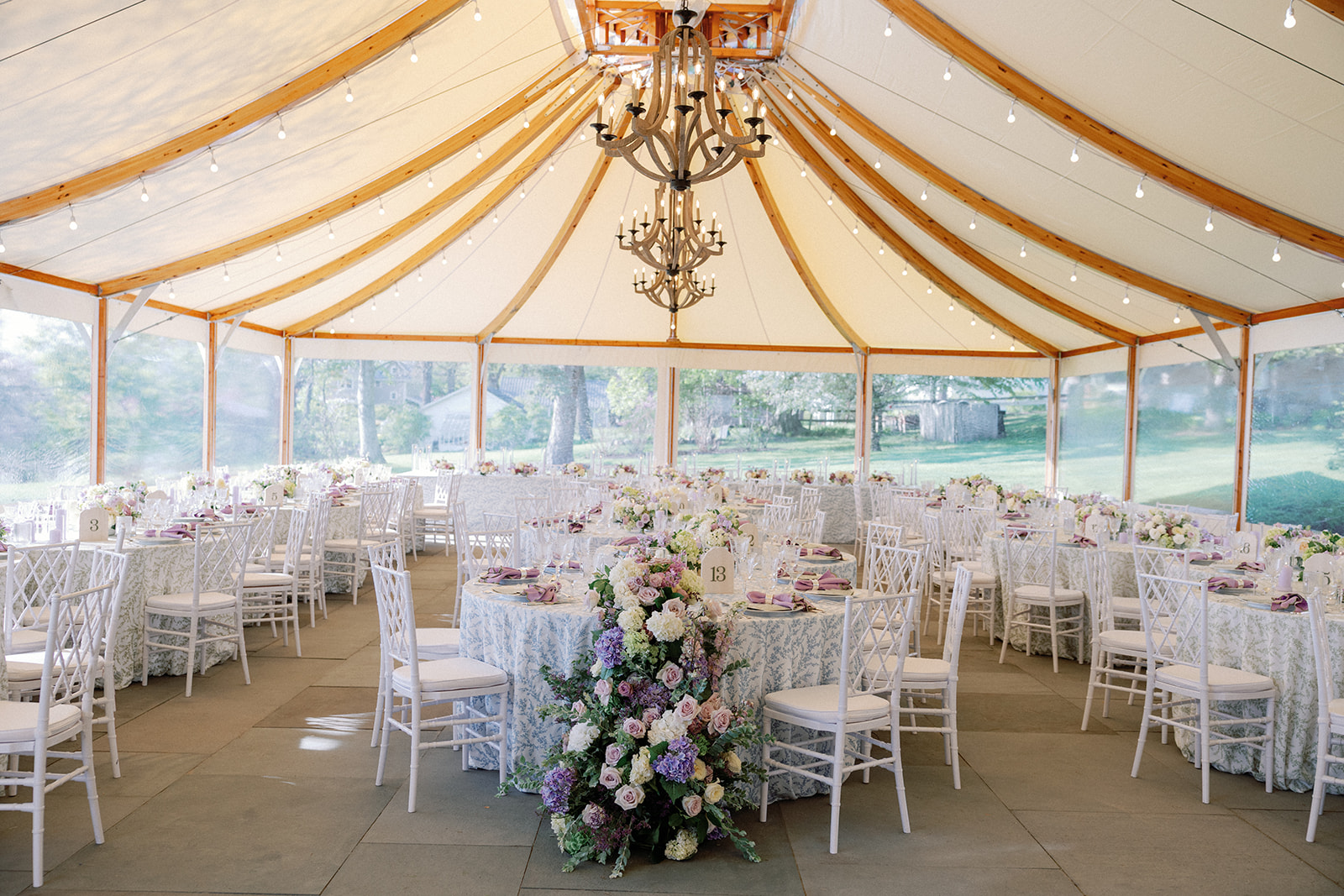 Wide interior view of a tented reception space with a peaked canvas ceiling, wooden beams, two tiered wood chandeliers, and string lights, with round tables set in pastel linens in the background.
