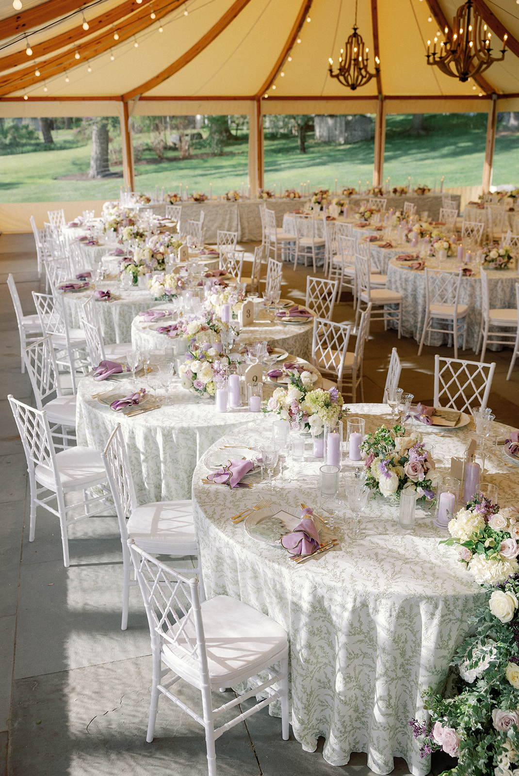 Rows of round reception tables inside a tent, set with pale blue floral-print linens and lavender napkins, with wooden chandeliers and string lights strung overhead.
