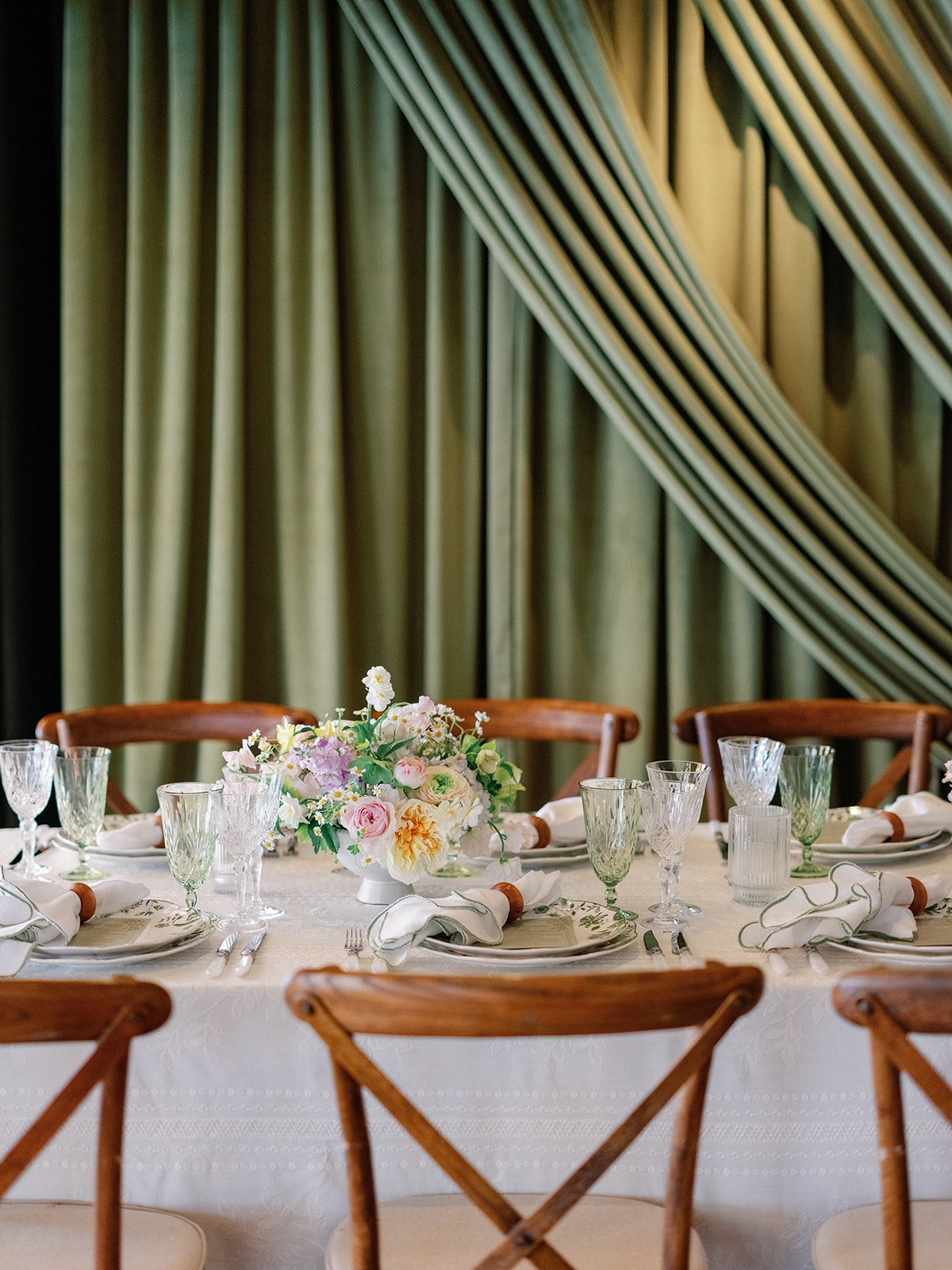Round table place setting with a pastel floral centerpiece in pink, yellow, and white, green and crystal glassware, and green-patterned china against a draped sage curtain.