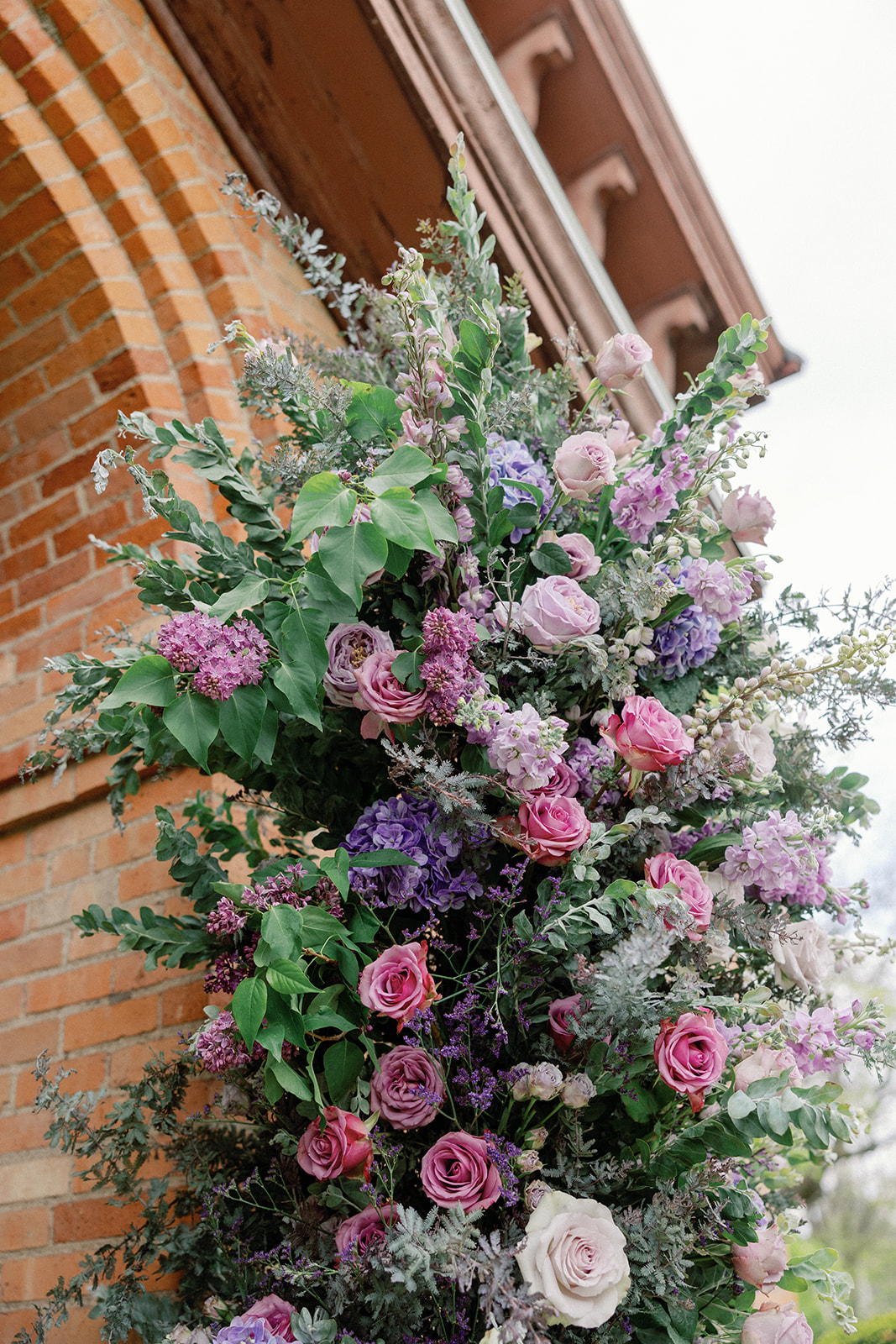 Close-up of a lush floral arrangement in pink, purple, and lavender tones cascading against an arched brick doorway.